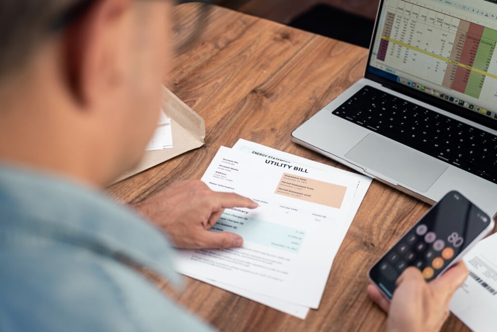 Man reviewing utility bills at home while using a smartphone calculator. Personal finance management and budgeting concept. Ideal for household expenses and tax calculation themes